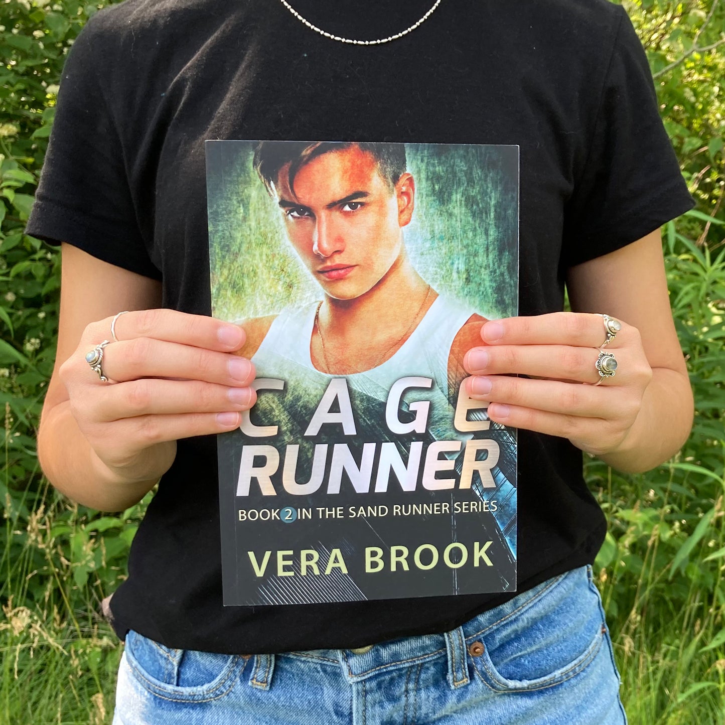 A teenage girl holds a paperback of Cage Runner, Book 2 in the Sand Runner Series by Vera Brook. It's a summer day. The girl is wearing a black tshirt and jeans and silver rings on her fingers. A very green forest is behind her.