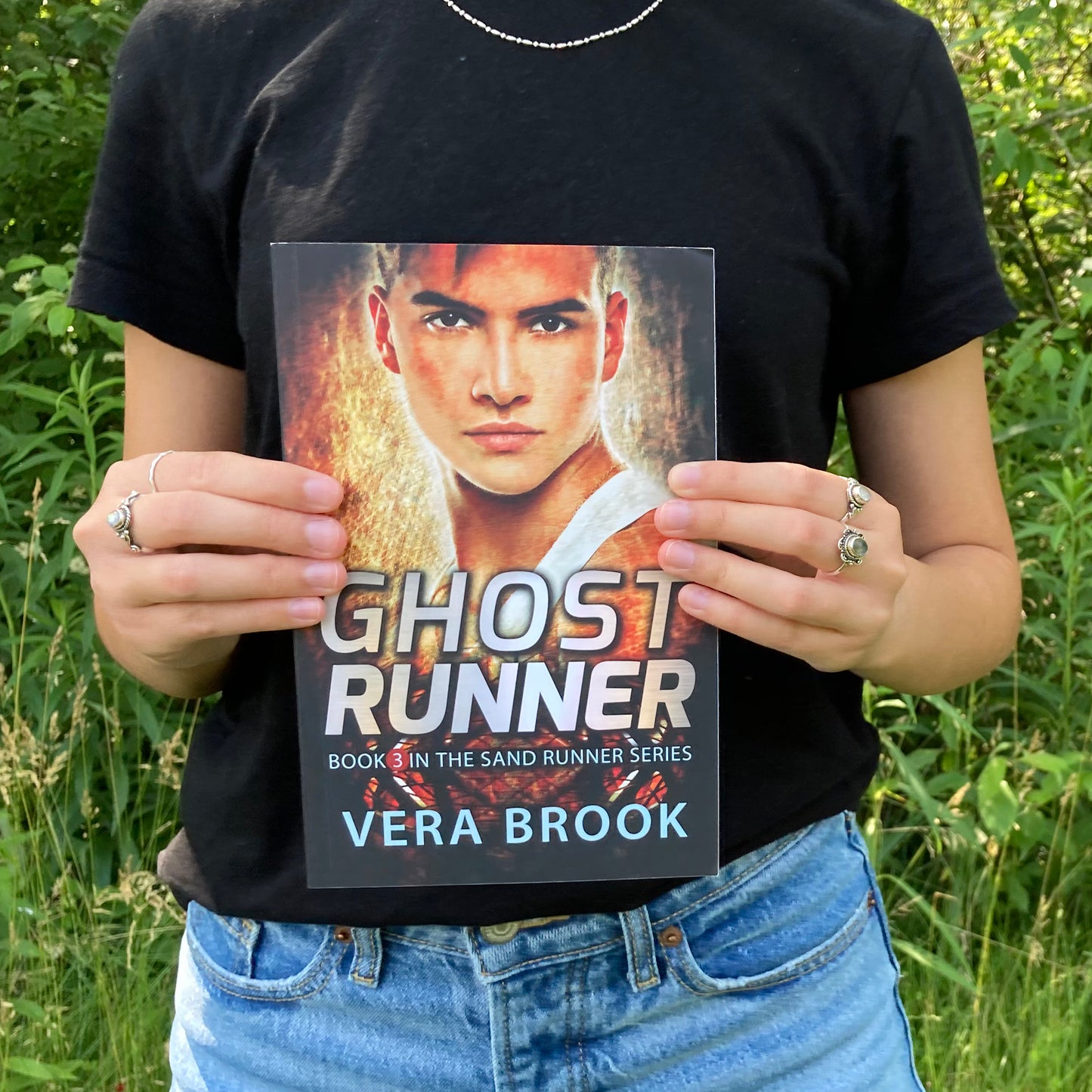 A teenage girl holds a paperback of Ghost Runner, Book 3 in the Sand Runner Series by Vera Brook. It's a summer day. The girl is wearing a black tshirt and jeans and silver rings on her fingers. A very green forest is behind her.