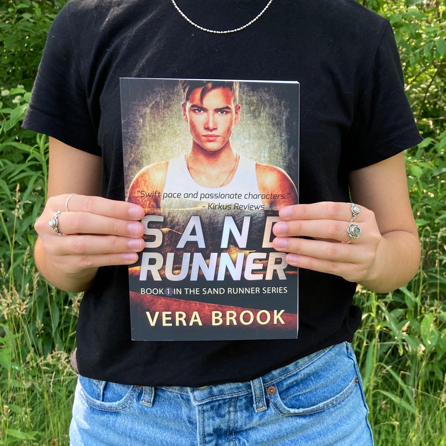 A teenage girl holds a paperback of Sand Runner, Book 1 in the Sand Runner Series by Vera Brook. It's a summer day. The girl is wearing a black tshirt and jeans and silver rings on her fingers. A very green forest is behind her.