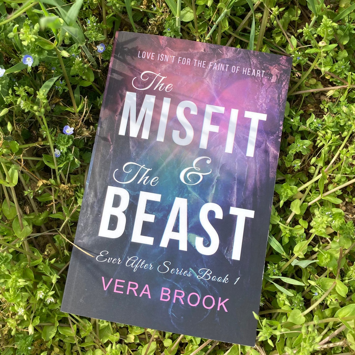 Paperback of The Misfit and The Beast by Vera Brook, lying flat on the green grass on a sunny summer day. The cover is purple, with the title in bold, white letters.