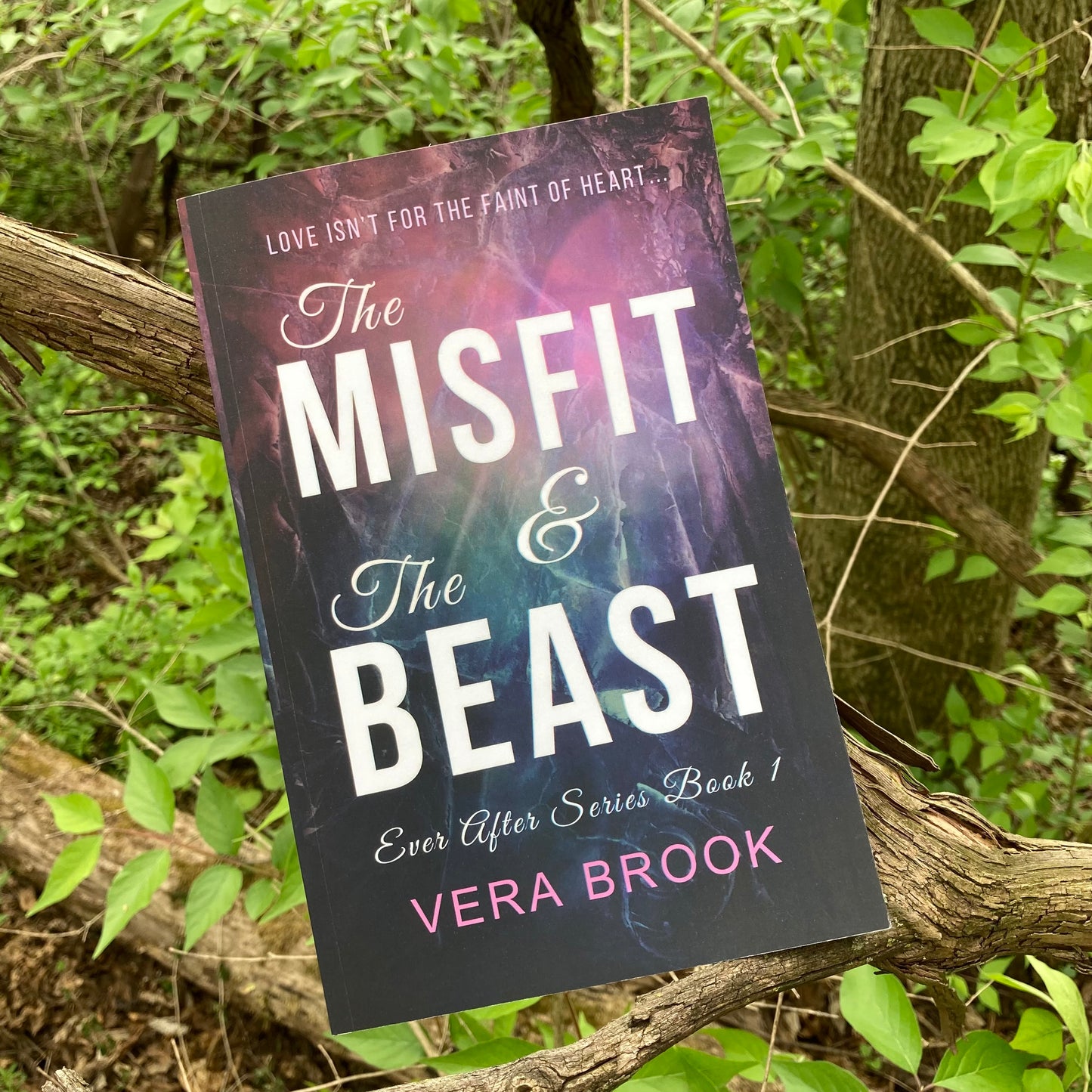 A paperback of The Misfit and The Beast, a young adult romance about first love and high school theater by Vera Brook. The book sits on a branch of a tree, with luscious green leaves all around it.