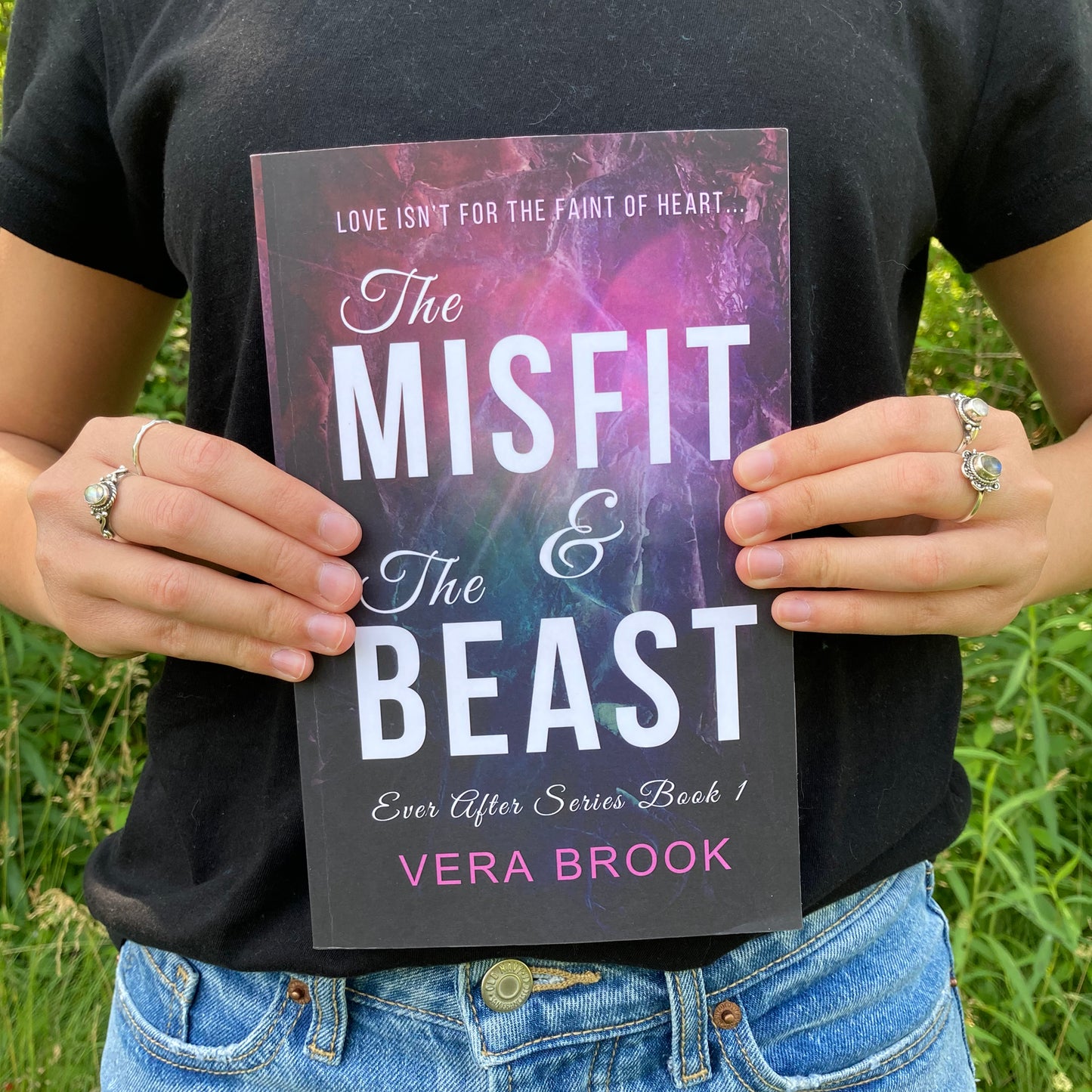 A teenage girl holds a paperback of The Misfit and The Beast by Vera Brook in front of her. She is wearing silver rings, a black tshirt, and jeans. She is standing outside on a summer day. The background is luscious green trees, bushes, and wildflowers.